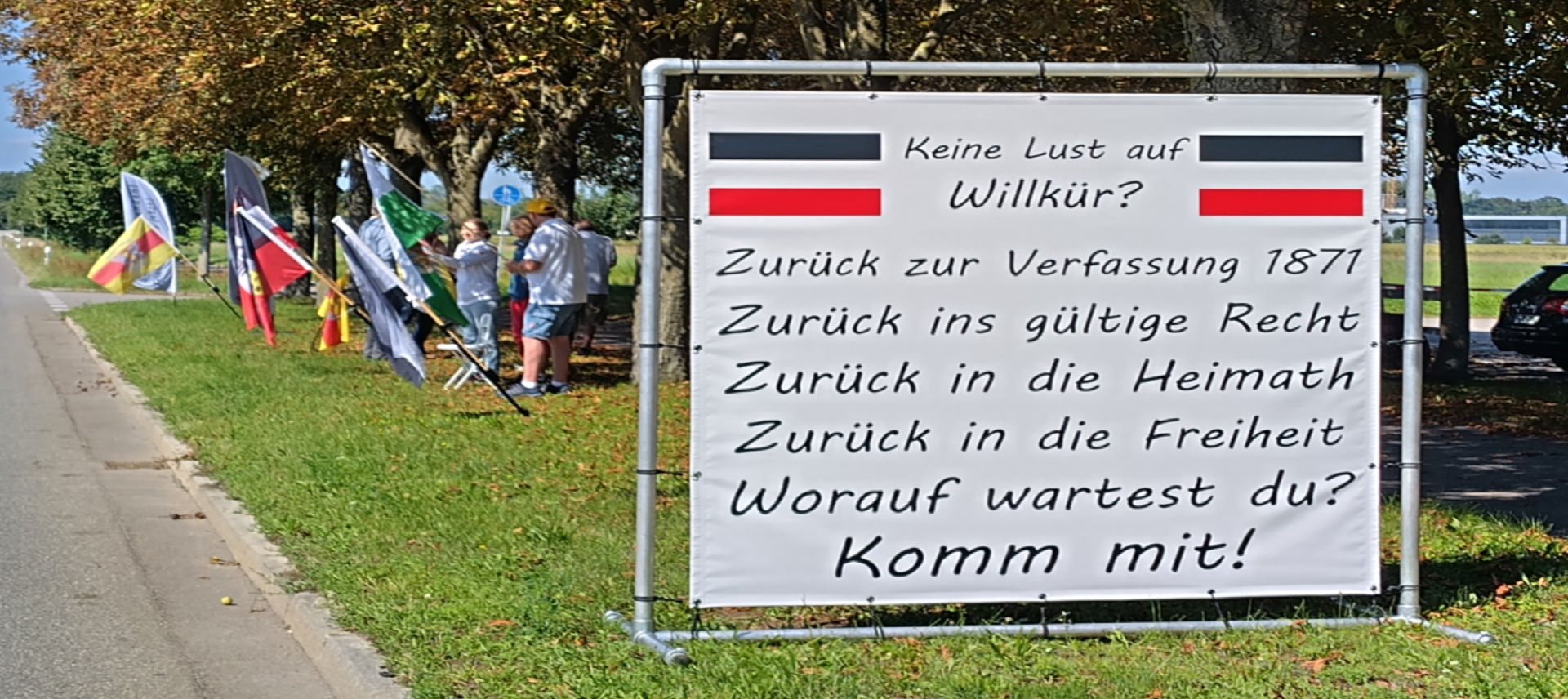 Menschen stehen auf einem Grünstreifen zwischen einer Straße und einem Radweg. Sie halten verschiedene Fahnen, darunter die Reichsflagge. Im Vordergrund steht ein großes Transparent. Unter zwei Reichsfahnen steht: Keine Lust auf Willkür? Zurück zur Verfassung 1871 Zurück ins gültige Recht Zurück in die Heimath Zurück in die Freiheit Worauf wartest du? Komm mit!