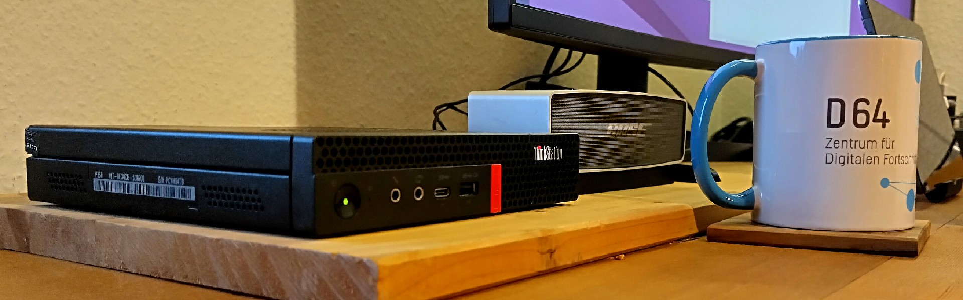 Foto des kleinen, schwarzen Rechners der Marke Lenovo. Daneben sieht man einen kleinen Bose-Lautsprecher und den untersten Rand des Monitors. Auch eine Tasse ist am rechten Bildrand zu sehen. Insgesamt ein typisch gestelltes Schreibtisch-Setting.