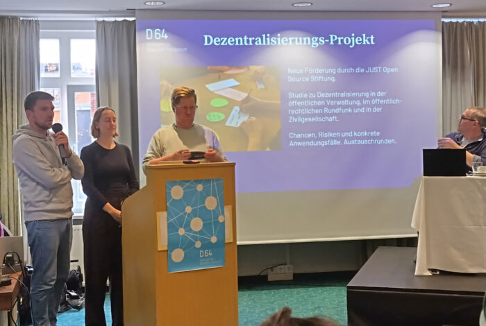 Zwei Männer und eine Frau reden zu einer Gruppe. Im Hintergrund eine Powerpoint-Folie.